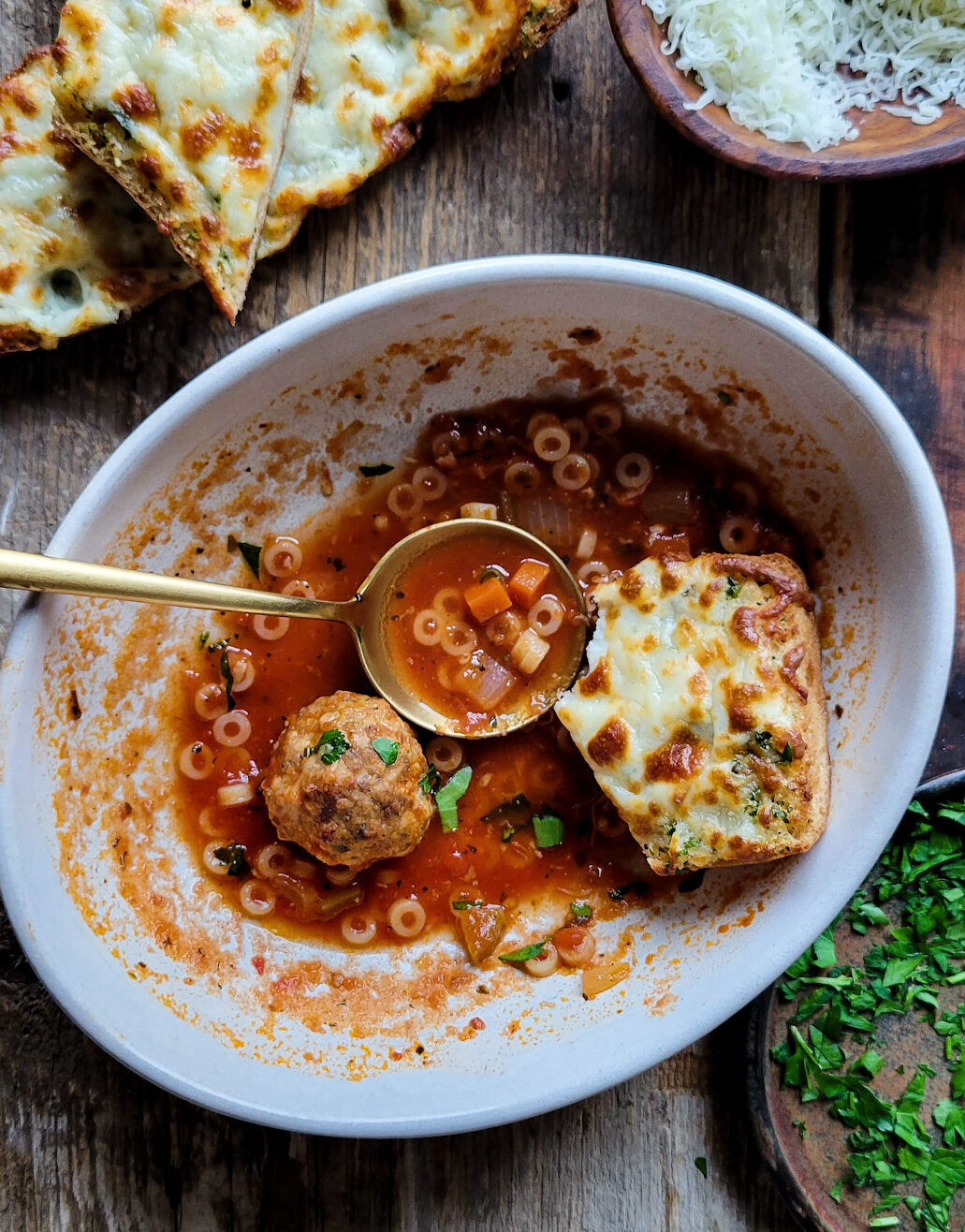 A bowl with just one meatball and a bit of soup left in it, with a spoon and half eaten piece of garlic bread are in the bowl as well, and more garlic bread slices are to the side of the bowl.