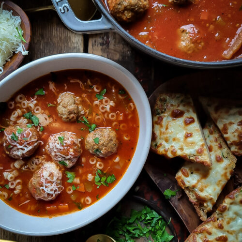 A pot of meatball soup is accompanied with a bowl of the soup, as well as slices of cheesy garlic bread