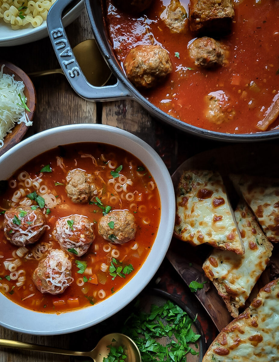 A pot of meatball soup is accompanied with a bowl of the soup, as well as slices of cheesy garlic bread