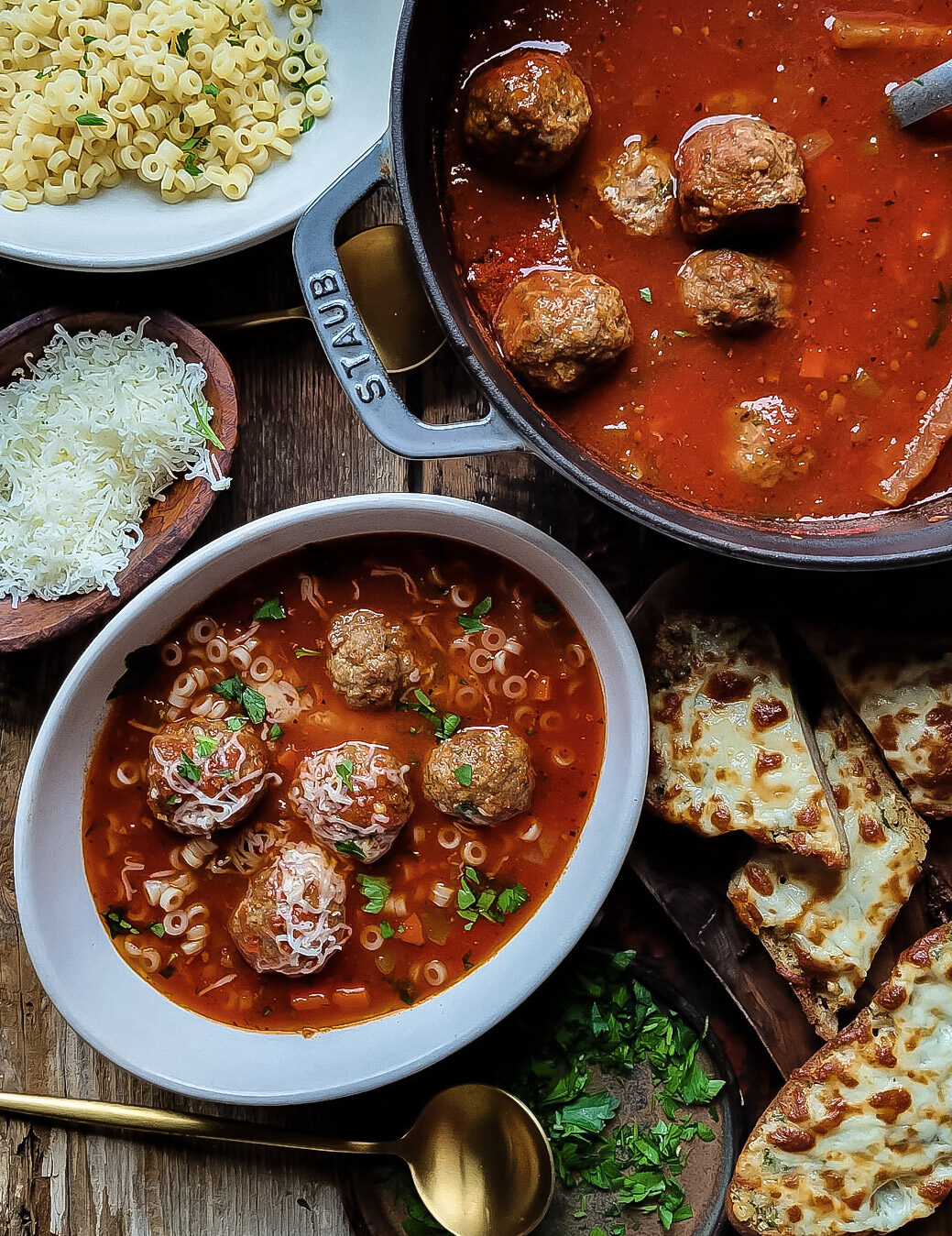 A pot of meatball soup is surrounded with bowls for the soup, as well as grated cheese, and cheesy garlic bread.