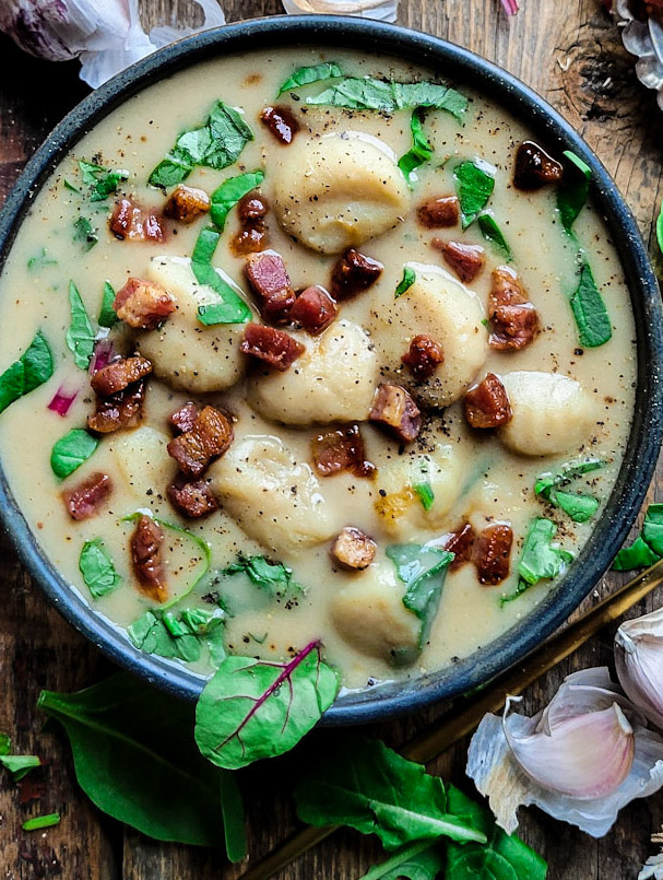 A bowl of comforting roasted garlic and gnocchi soup, topped with pancetta and greens
