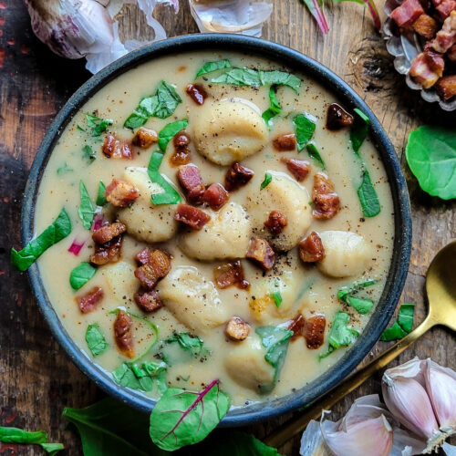 A bowl of hearty roasted garlic and gnocchi soup with greens and pancetta. Garlic cloes and spinach are scattered around the bowl.