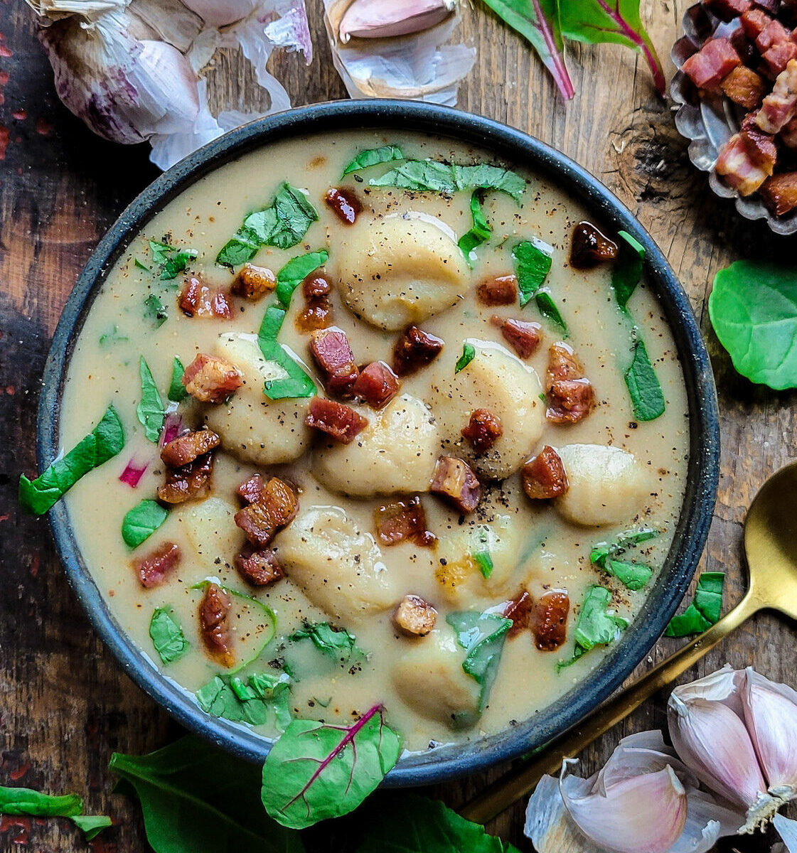 A bowl of hearty roasted garlic and gnocchi soup with greens and pancetta. Garlic cloes and spinach are scattered around the bowl.