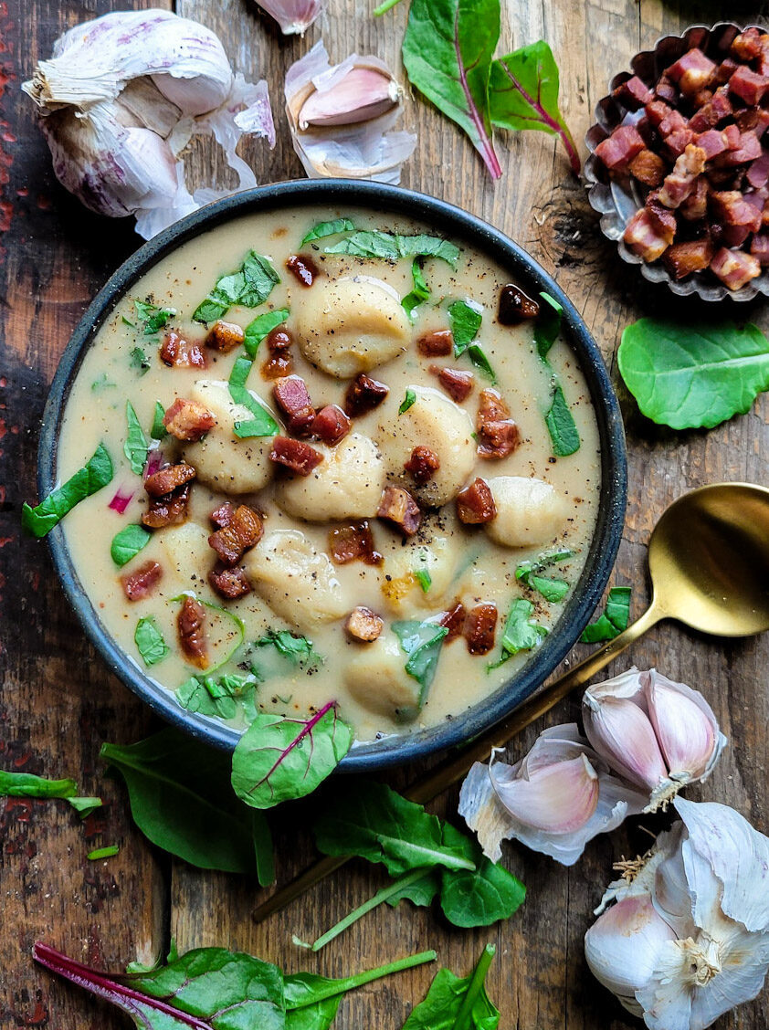 A bowl of hearty roasted garlic and gnocchi soup with greens and pancetta. Garlic cloes and spinach are scattered around the bowl.