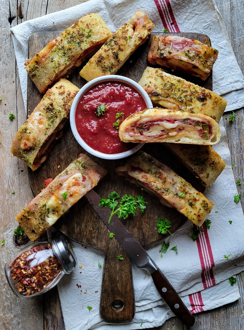 A cutting board with sliced up Stromboli or rolled pizza, with harissa marinara dipping sauce.