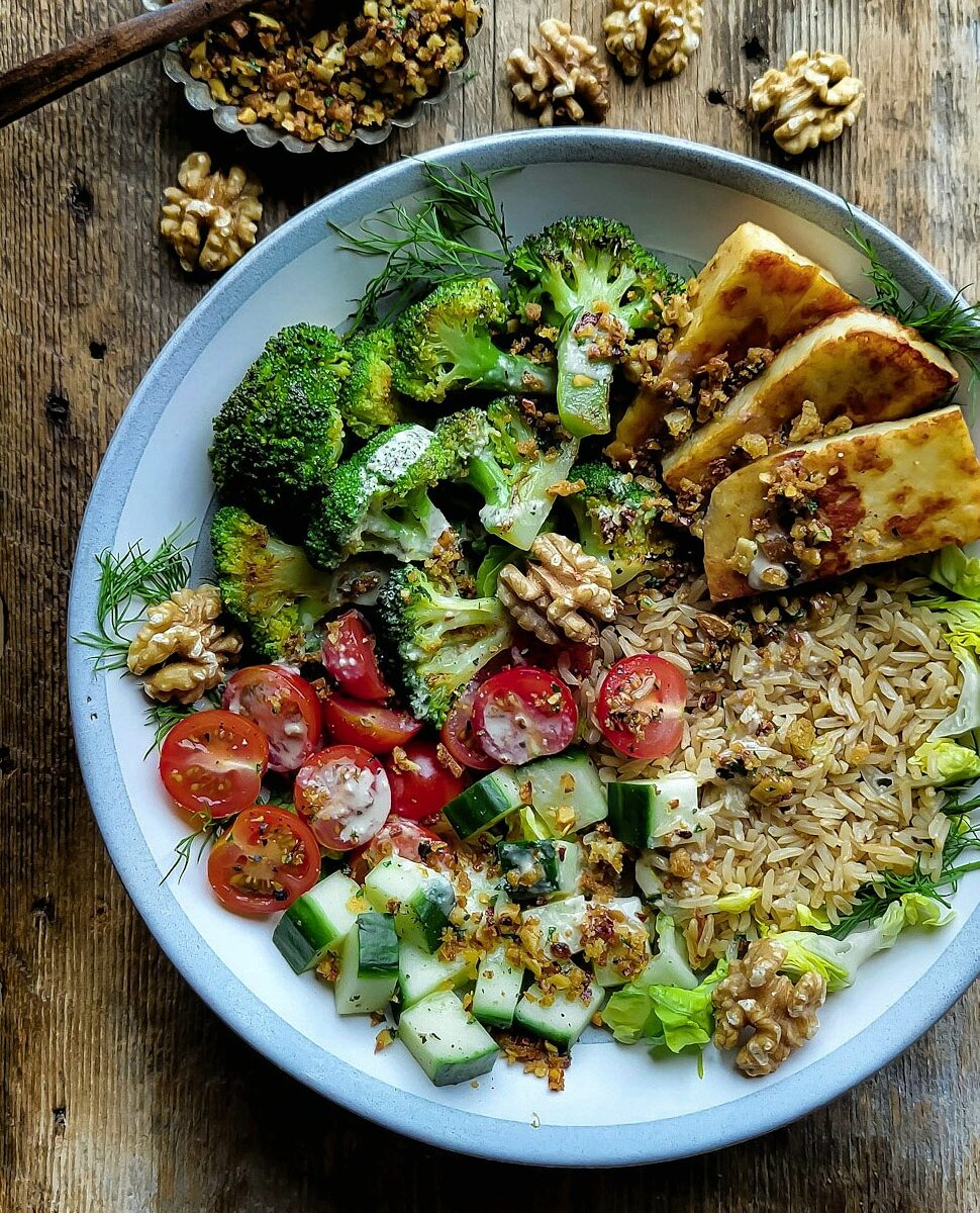 Brown Rice and Broccoli bowl filled with pan fried halloumi, fresh tomatoes and cucumbers, brown rice and roasted broccoli. Fresh walnuts and a small bowl of walnut pangrattato are to the side.