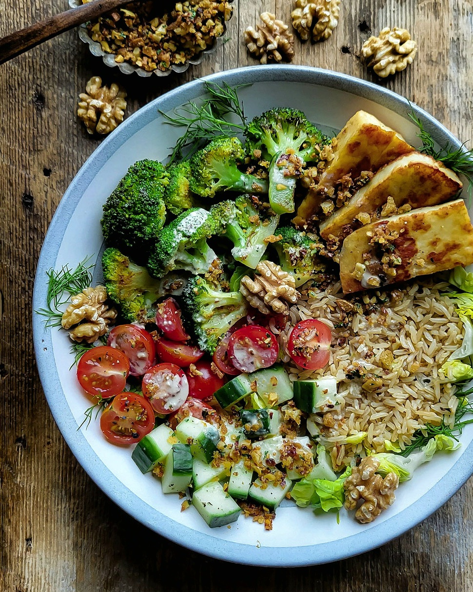 Brown Rice and Broccoli bowl filled with pan fried halloumi, fresh tomatoes and cucumbers, brown rice and roasted broccoli. Fresh walnuts and a small bowl of walnut pangrattato are to the side.