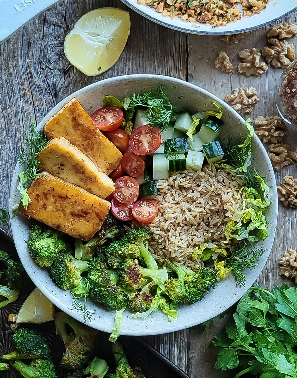 Brown Rice and Broccoli bowl filled with pan fried halloumi, fresh tomatoes and cucumbers, brown rice and roasted broccoli. Lemon wedge and walnuts are scattered around the bowl.