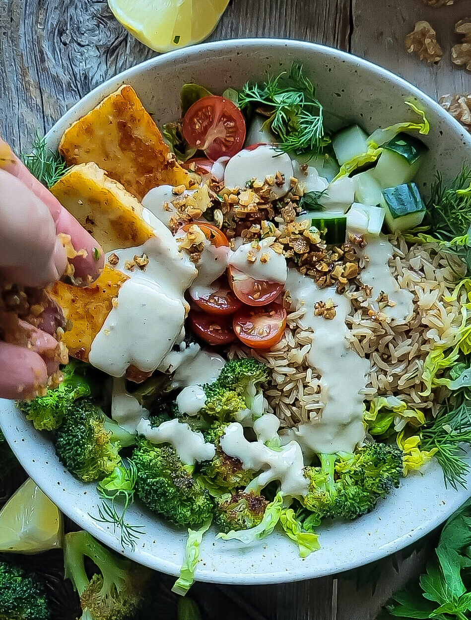 Brown Rice and Broccoli bowl filled with pan fried halloumi, fresh tomatoes and cucumbers, brown rice and roasted broccoli. A hand is shown sprinkling walnut pangrattato over everything.