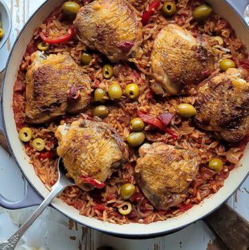 A skillet sitting on a cutting board is filled with spiced rice and chicken thighs, along with roasted peppers, olives and chorizo sausage.
