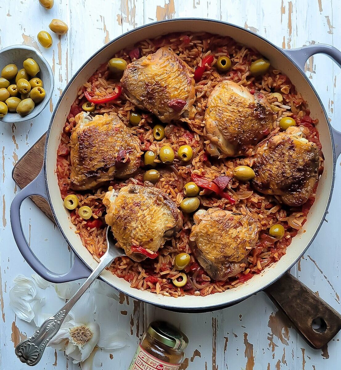 A skillet sitting on a cutting board is filled with spiced rice and chicken thighs, along with roasted peppers, olives and chorizo sausage. a bowl of olives and garlic cloves are scattered around the cutting board.