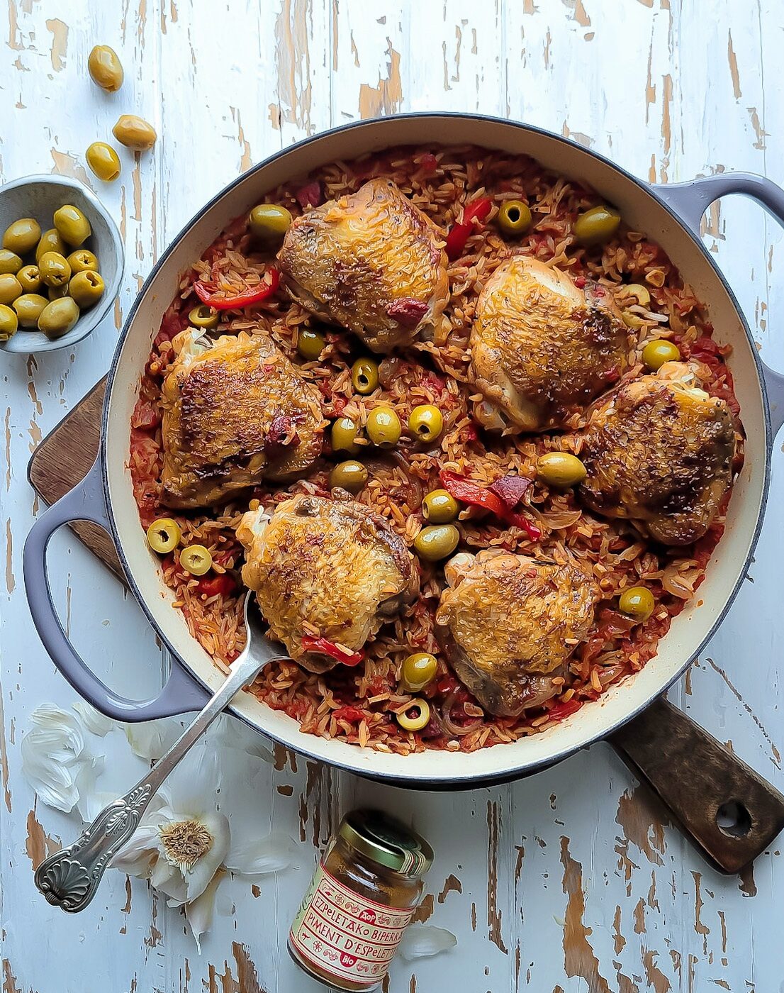 A skillet sitting on a cutting board is filled with spiced rice and chicken thighs, along with roasted peppers, olives and chorizo sausage. a bowl of olives and garlic cloves are scattered around the cutting board.