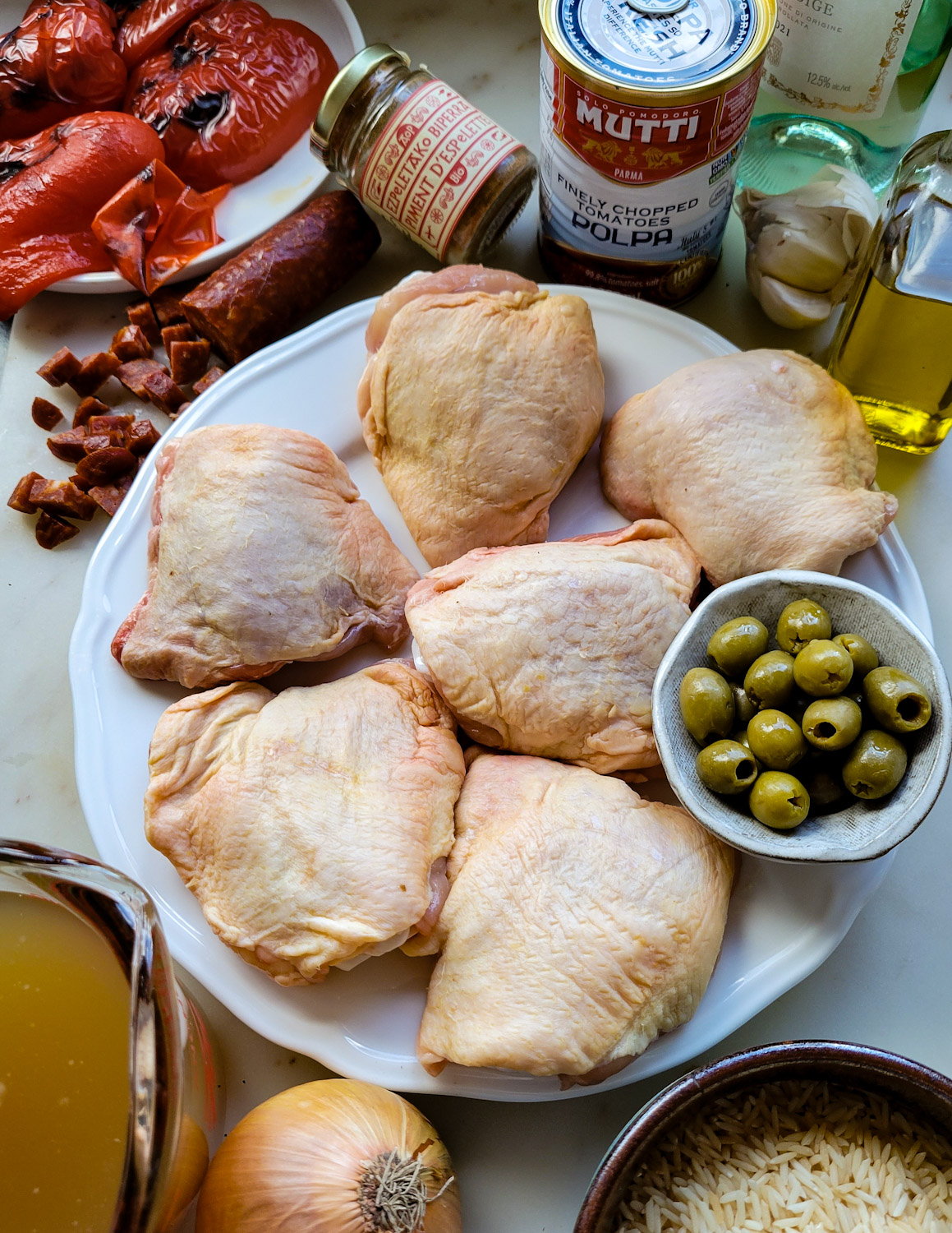 Ingredients for making Basque Chicken and Rice are gathered together on a counter. Roasted red peppers, espelette pepper, crushed tomatoes, olive oil, garlic, chicken thighs, olives, rice, onion and stock.