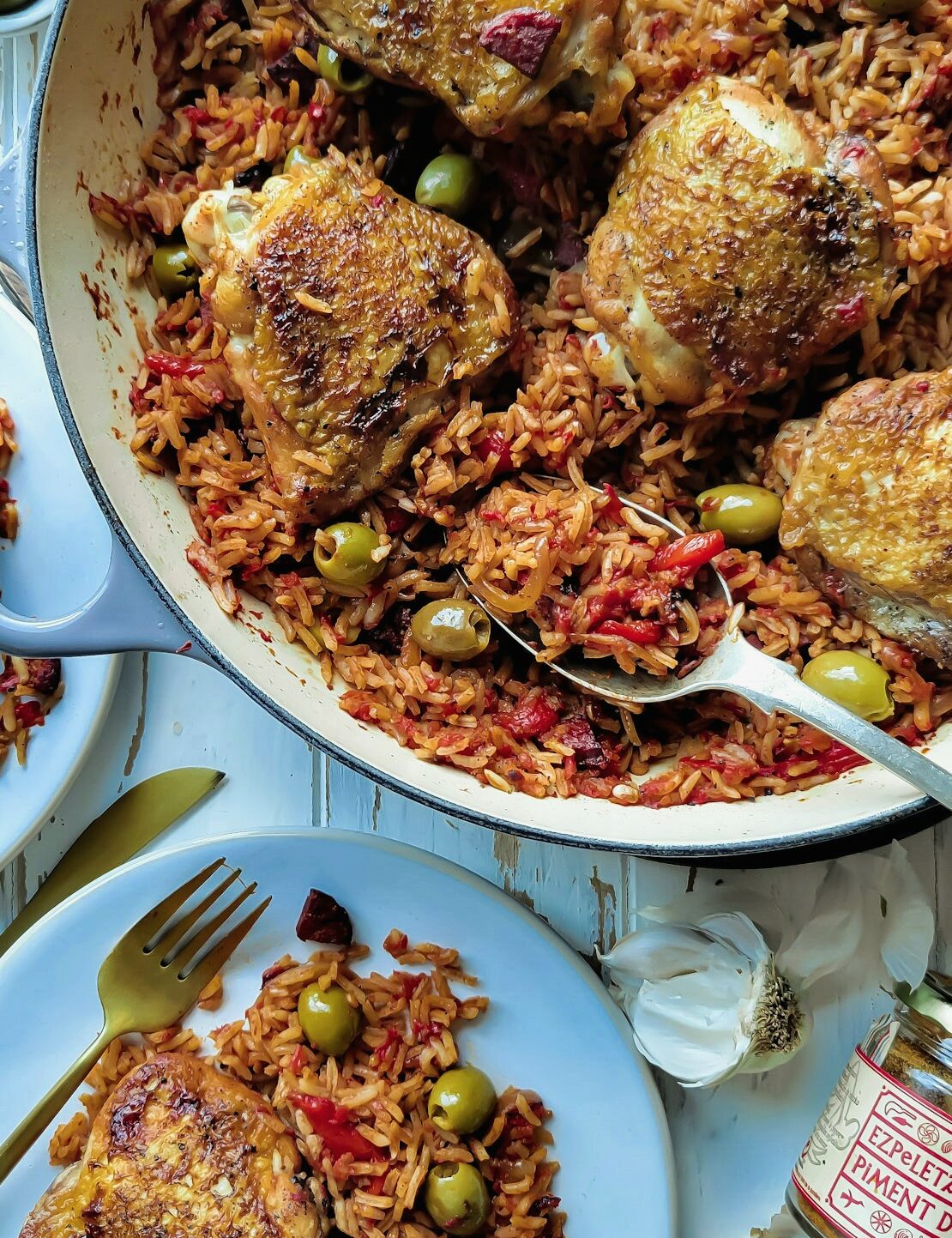 A skillet of Basque Chicken and Rice with a plate containing one serving nearby. A serving spoon is nestled in the skillet.