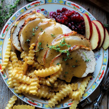 A plate with a sumptuous layered hot turkey sandwich, fries, and lingonberry preserves and apple slices. Gravy and fresh thyme leaves garnish the meal.