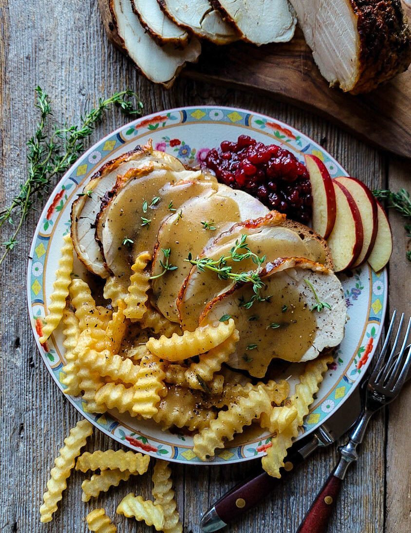 A plate with a sumptuous layered hot turkey sandwich, fries, and lingonberry preserves and apple slices. Gravy and fresh thyme leaves garnish the meal.