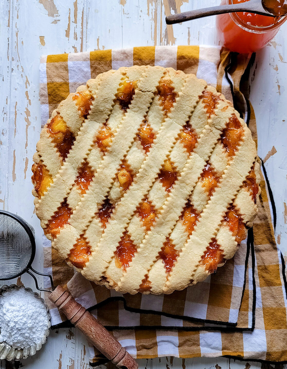 A baked Marmalade Ricotta Crostata is sitting on a tea towel, with marmalade jam and spoon to the side, as well as icing sugar and a mesh strainer.