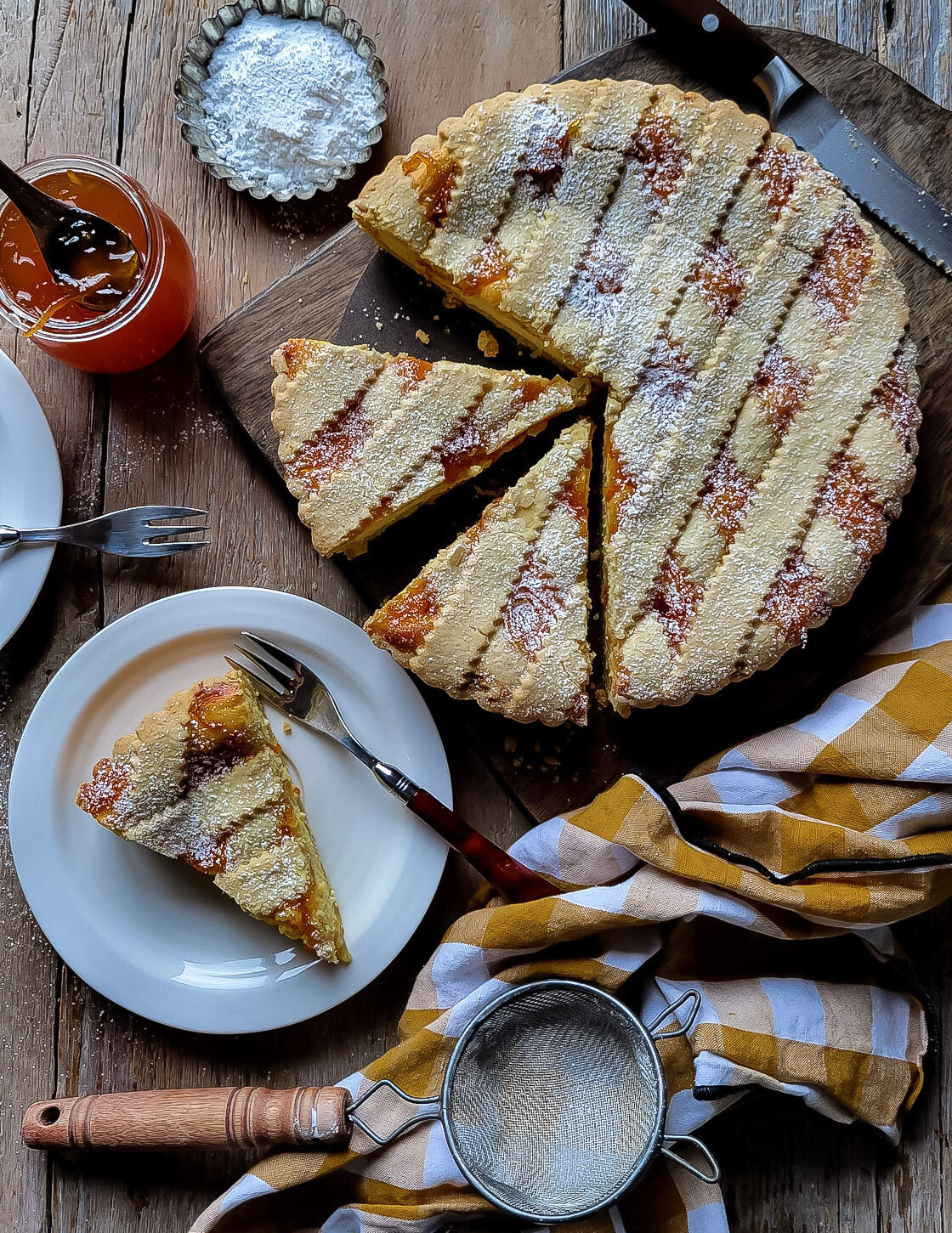 A tablescape of baked Marmalade Ricotta Crostata on a cutting board, with a knife, a jar of marmalade and tea towel surrounding it. A plate with a slice, as well as icing sugar and a mesh strainer are on the side.