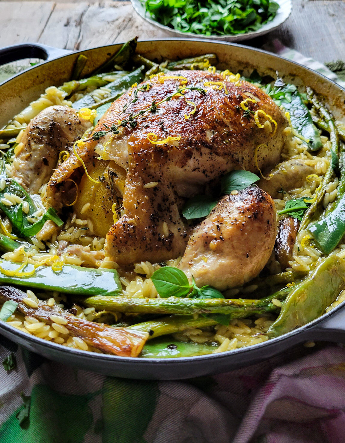 A pot filled with Spring Chicken and Orzo and charred vegetables. A plate of spring greens are in the background.