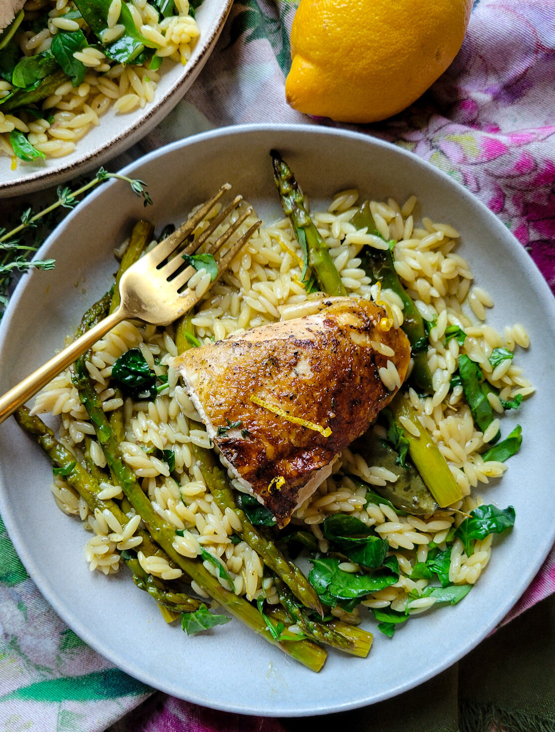 A plate with a serving of One Pot Spring Chicken and Orzo with spring vegetables, a lemon and fresh thyme are to the side.