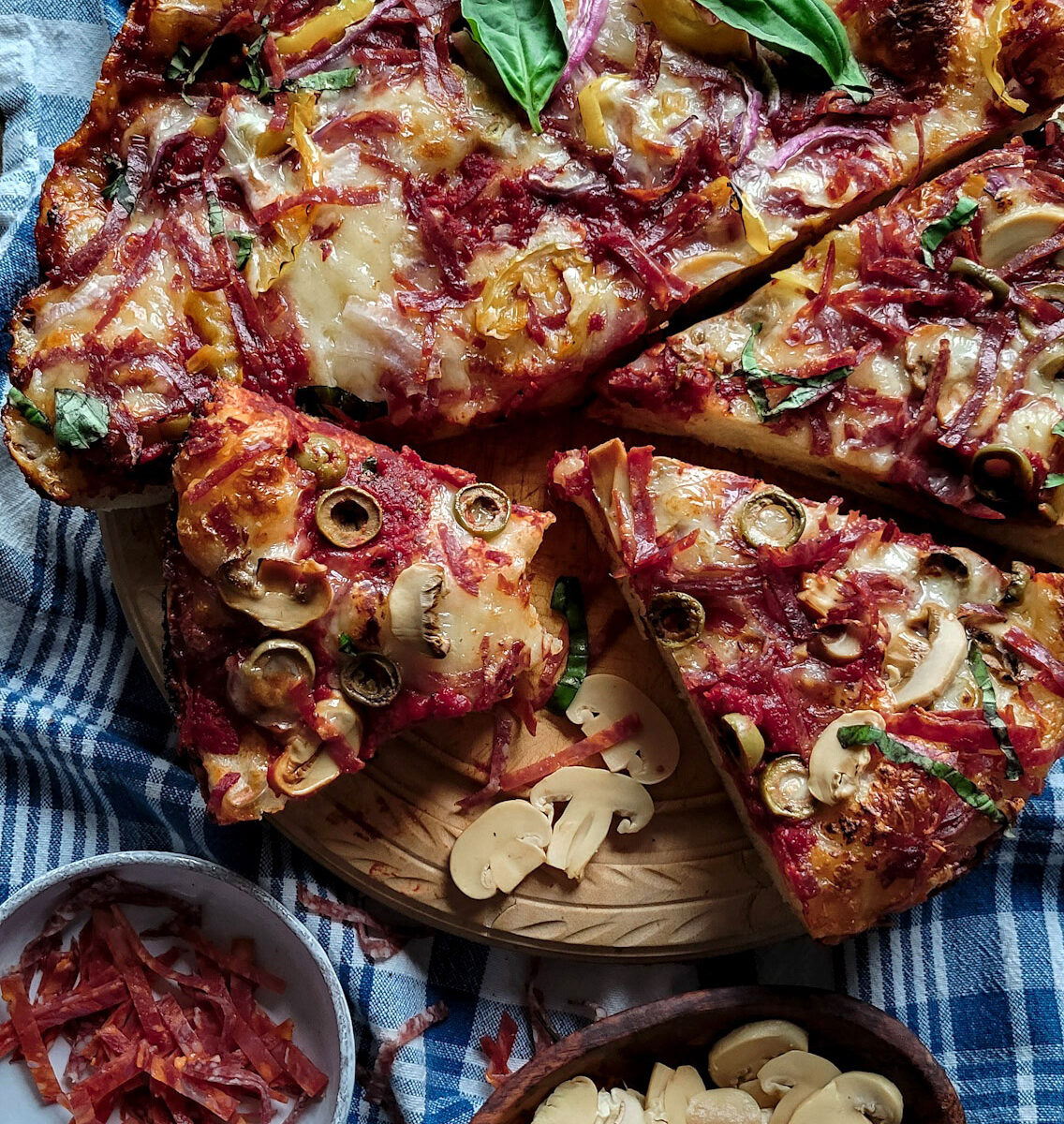 A sliced Windsor Style Skillet pizza on a cutting board, with thinly sliced pepperoni, and canned mushrooms to the side of the pizza.