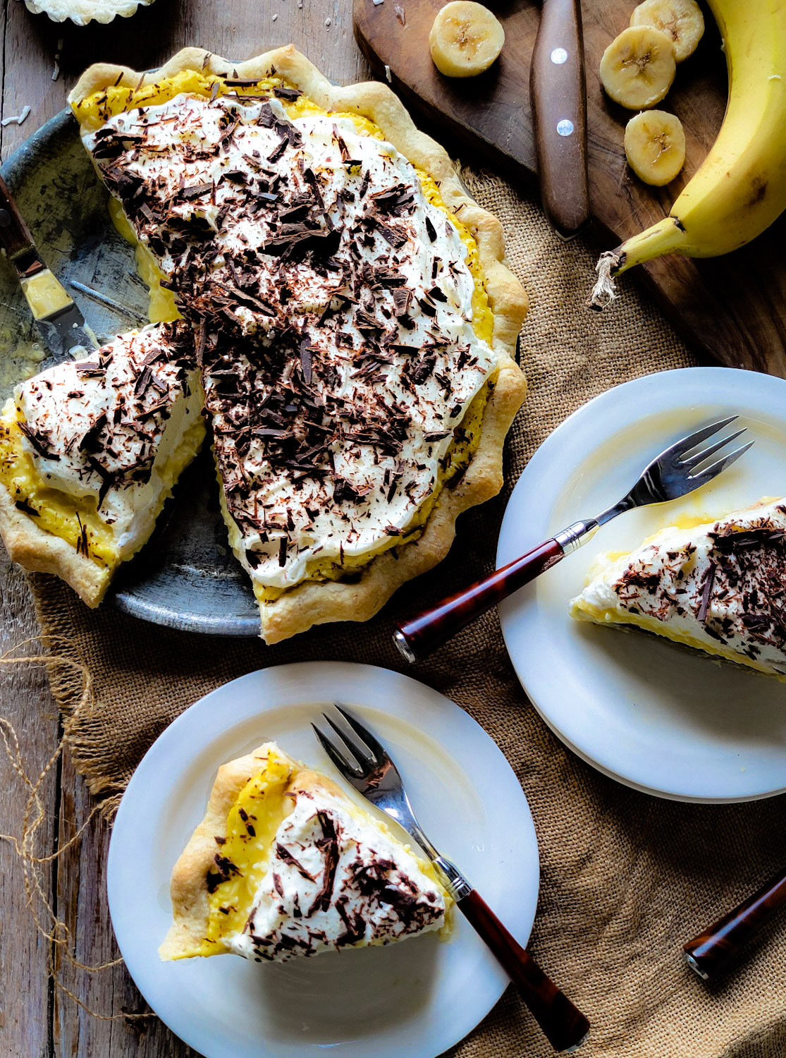 A full Black Bottom Banana Coconut Cream Pie, with two slices served up, and a sliced banana in the background.