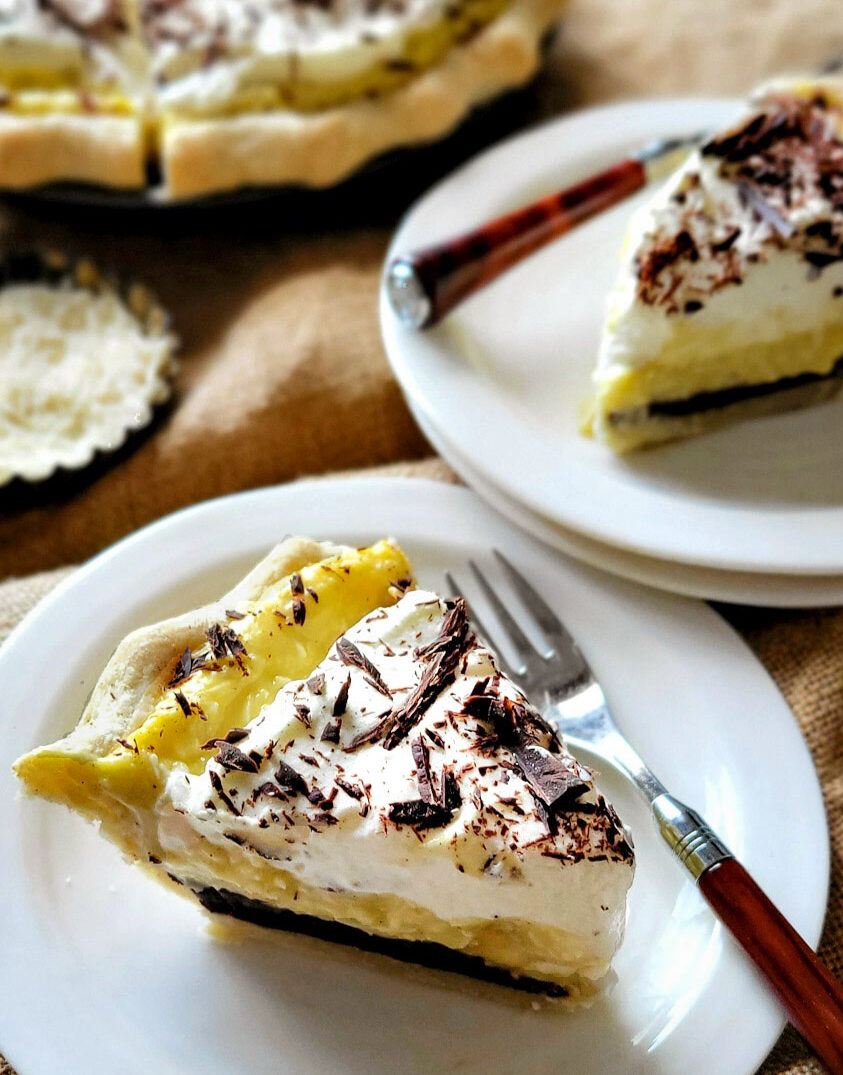 Close up of a slice of Black Bottom Banana Coconut Cream Pie. The rest of the pie, as well as another slice on a plate are in the background.