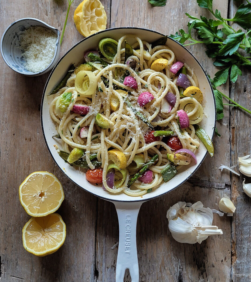 A skillet filled with colourful Lemony Pasta Primavera. IT is surrounded by parsley, garlic cloves, a bowl of grated parmesan cheese, and juiced lemons.