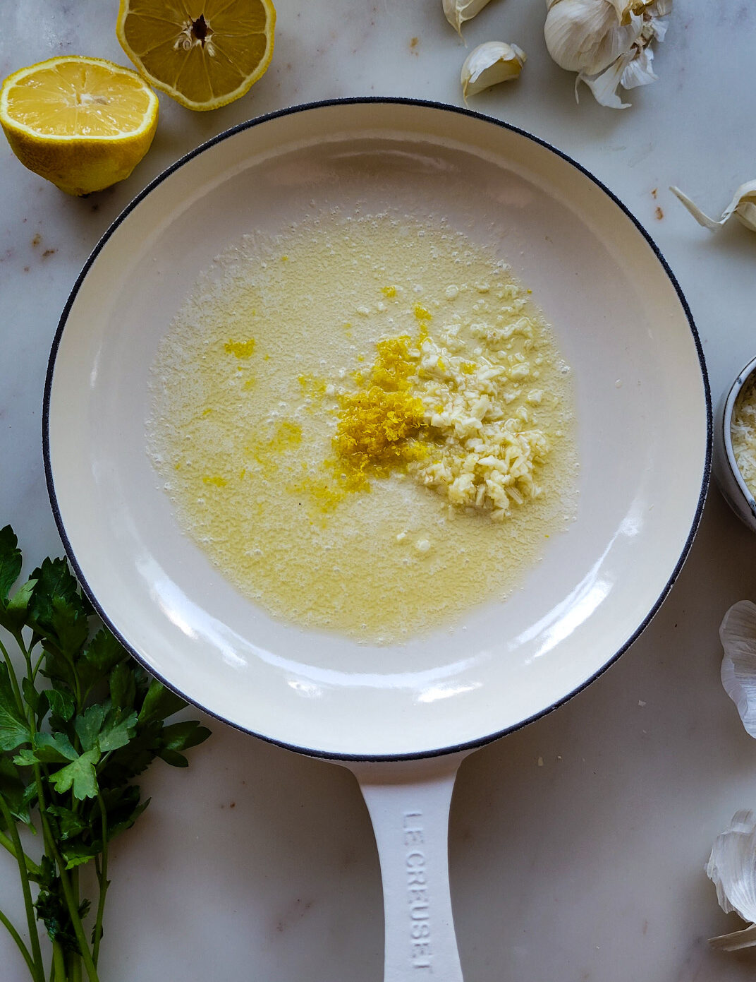 A skillet with melted butter, minced garlic and lemon zest ready for pasta to be added to it. There is a sliced lemon, and garlic cloves, as well as parsley in the background.