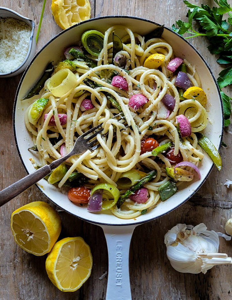 A skillet filled with colourful Lemony Pasta Primavera. IT is surrounded by parsley, garlic cloves, a bowl of grated parmesan cheese, and juiced lemons.