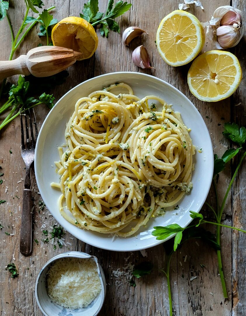 A plate of buttery, garlicky Lemon Pasta, surrounded by sliced lemon, garlic cloves, parmesan cheese and parsley.