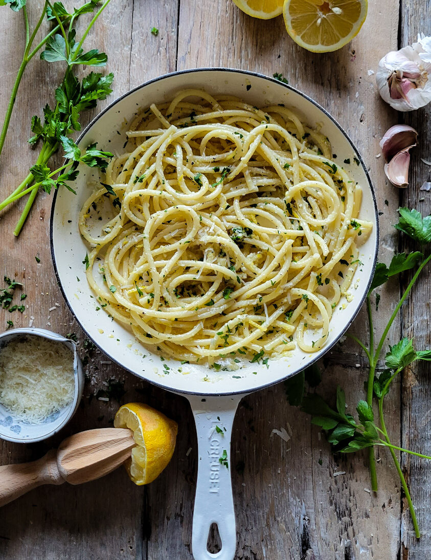 A skillet filled with colourful Lemon Pasta. It is surrounded by parsley, garlic cloves, a bowl of grated parmesan cheese, and juiced lemons.