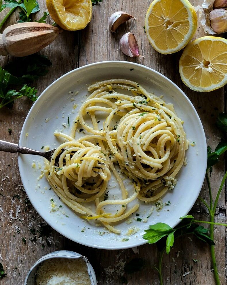 A plate of buttery, garlicky Lemon Pasta, surrounded by sliced lemon, garlic cloves, parmesan cheese and parsley.