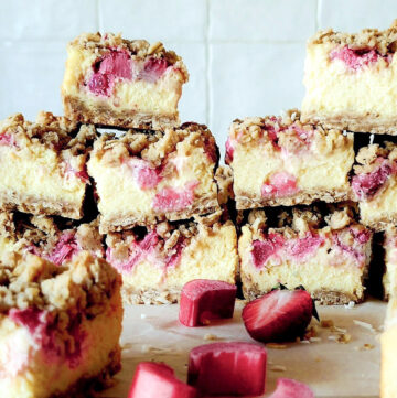 Strawberry Rhubarb Cheesecake squares cut and sitting on the counter surrounded by fresh strawberries and rhubarb.