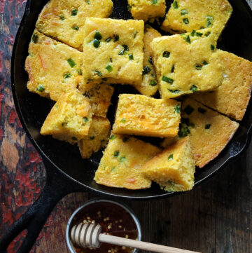 Jalapeno Cheddar Cornbread sliced up and sitting in an iron skillet with a jar of hot honey on the side.
