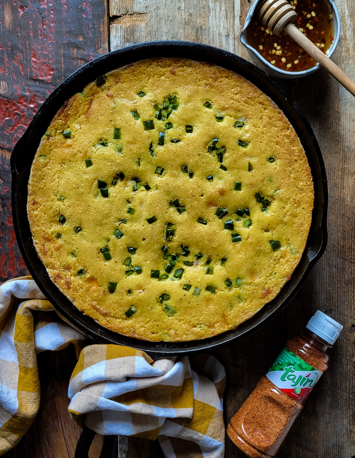 A skillet with a baked Jalapeno Cheddar Cornbread is sitting on a wooden counter, with hot honey and Tajín seasoning to the side.