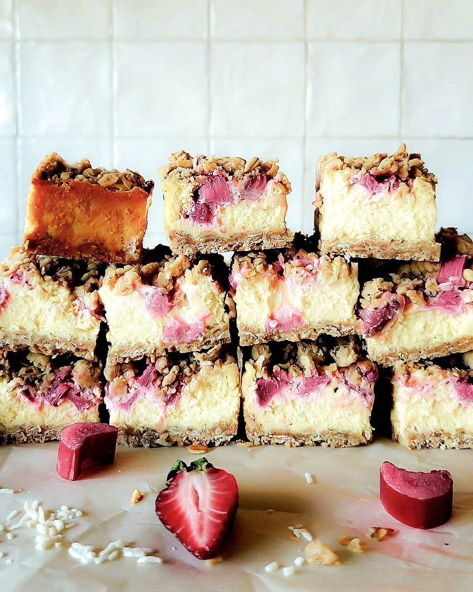 Strawberry Rhubarb Cheesecake squares cut and sitting on the counter surrounded by fresh strawberries and rhubarb.
