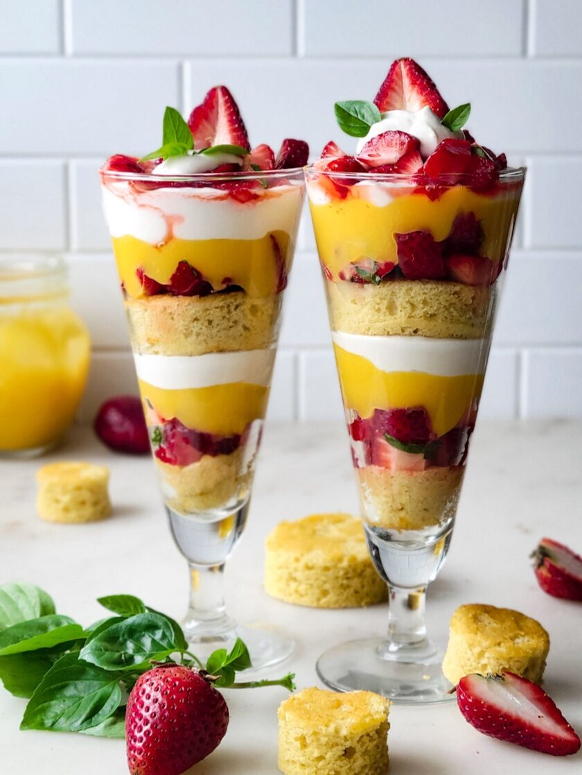Basil Strawberry Shortccake Parfait in two tall glasses, with berries and basil leaves scattered around them.