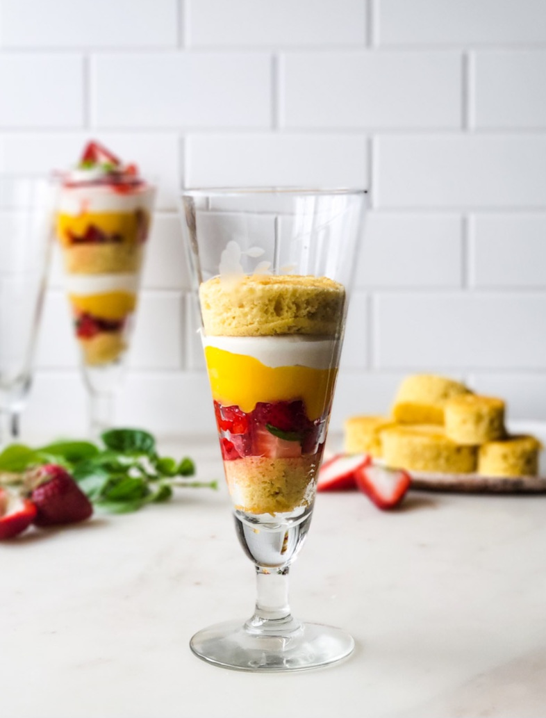 The making of a Basil Strawberry Shortcake Parfait. The layers of sponge cake, lemon curd and mascarpone whipped cream along with the berries are seen in the tall glass.