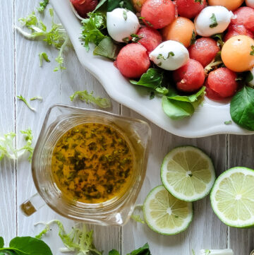 A serving platter filled with Agua Fresca Melon Salad, with the Hot Honey Lime Dressing to the side. Lime slices and salad greens are scattered about.