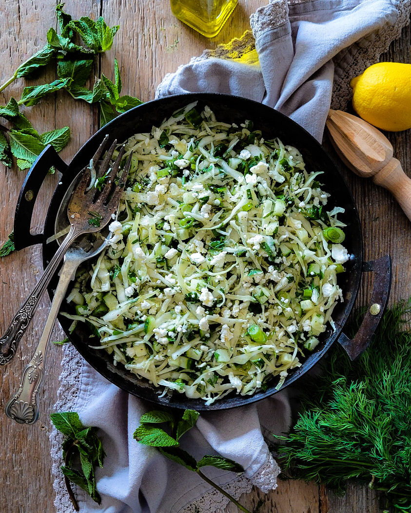 A serving dish filled with Cabbage, Mint and Feta Salad, with fresh mint leaves and dill scattered around it. A lemon and bottle of olive oil are in the background.