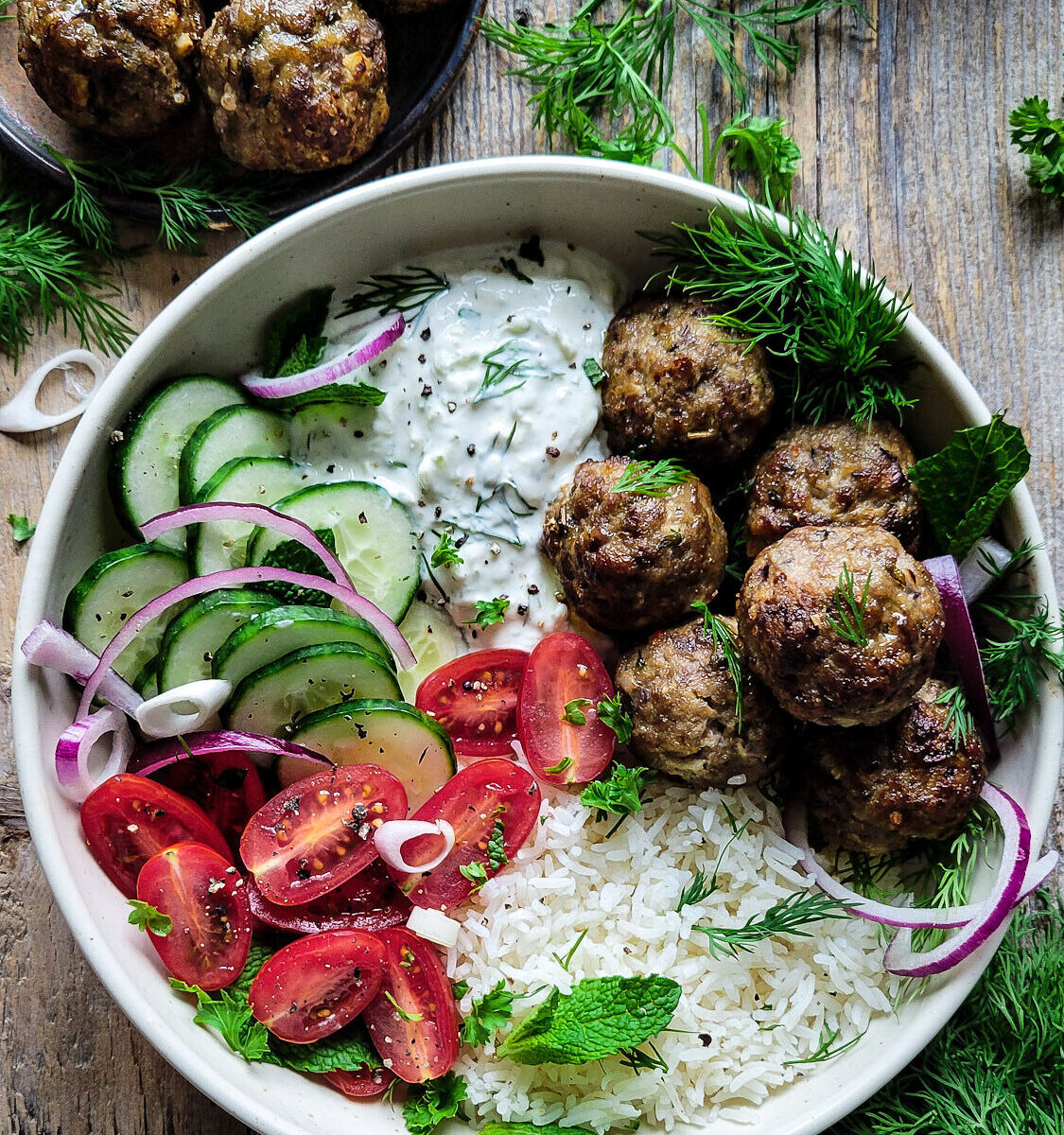 A Greek Bowl filled with Feta Stuffed Greek Meatballs, rice, Labneh Tzatziki, and sliced fresh cucumbers, tomatoes and onion. Fresh herbs are scatterred about the bowl.