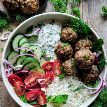 A Greek Bowl filled with Feta Stuffed Greek Meatballs, rice, Labneh Tzatziki, and sliced fresh cucumbers, tomatoes and onion. Fresh herbs are scatterred about the bowl.