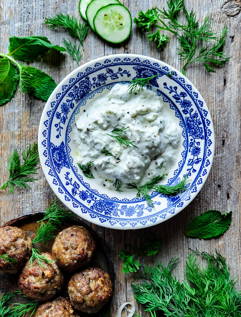 Feta Stuffed Greek Meatballs surrounding a bowl of Labneh Tzatziki. Cucumber slices, mint and dill surround everything.