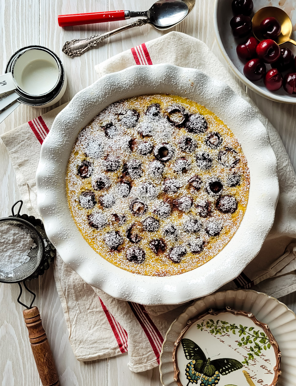 Cherry Clafoutis dusted with icing sugar, with a bowl of pitted cherries to the side.