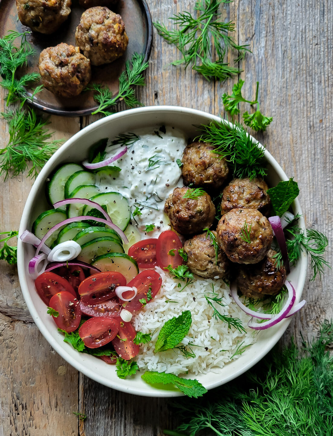 A bowl filled with Feta Stuffed Meatballs, Labneh Tzatziki, rice and fresh vegetables. A plate of more meatballs in in the background. Fresh herbs are scattered about.