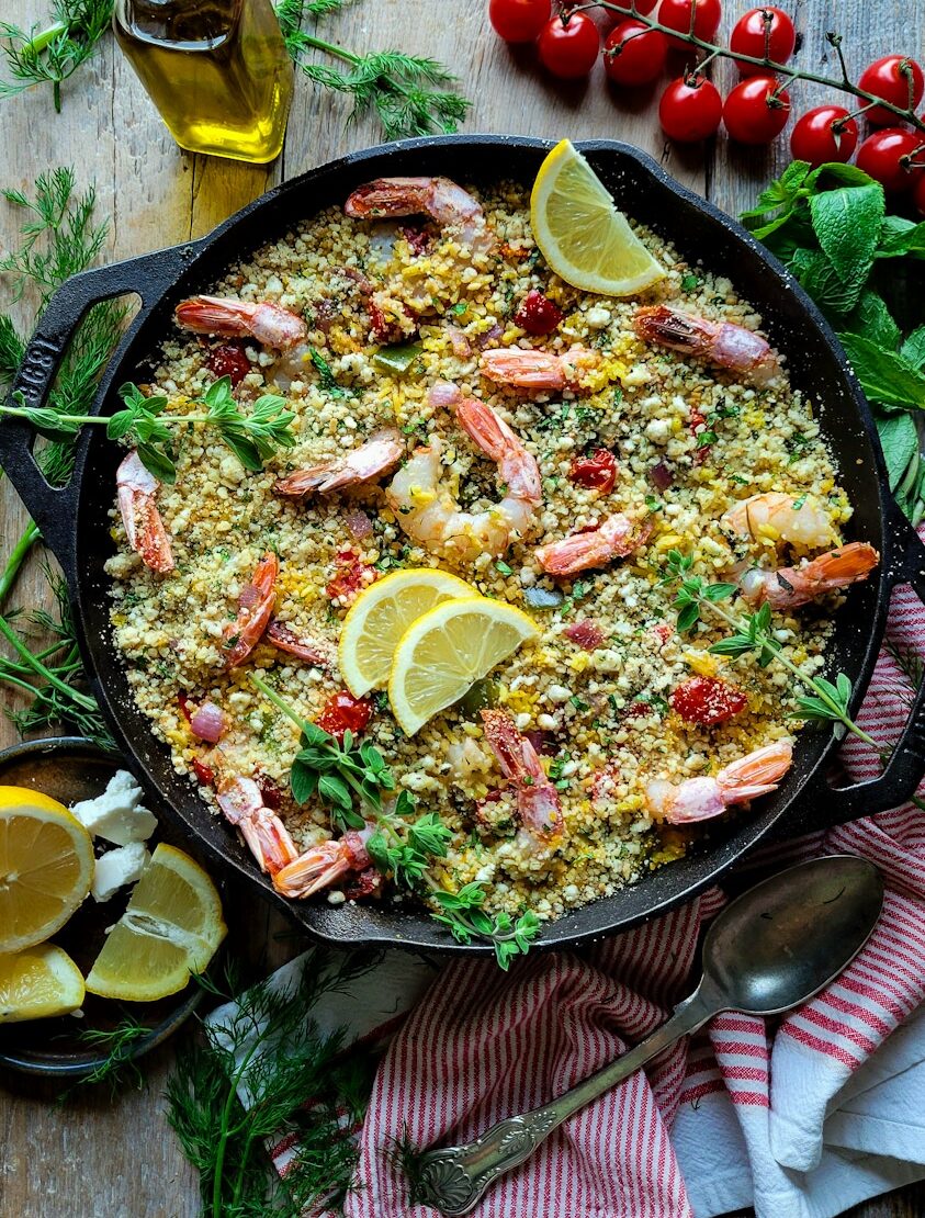 A skillet filled with Mediterranean Shrimp and Rice Bake, surrounded by cherry tomatoes, olive oil, fresh herbs and lemon slices.