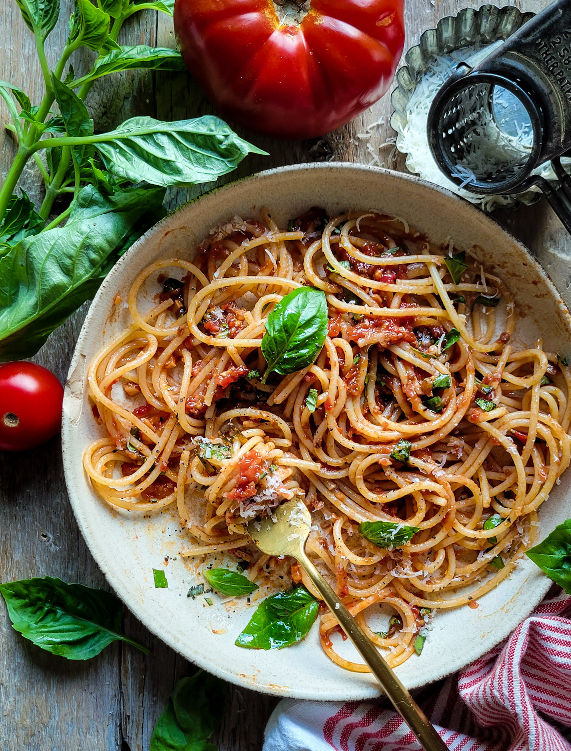 A bowl of Spaghetti with fresh Summer Sauce is surrounded by fresh basil, tomatoes and a cheese grater.
