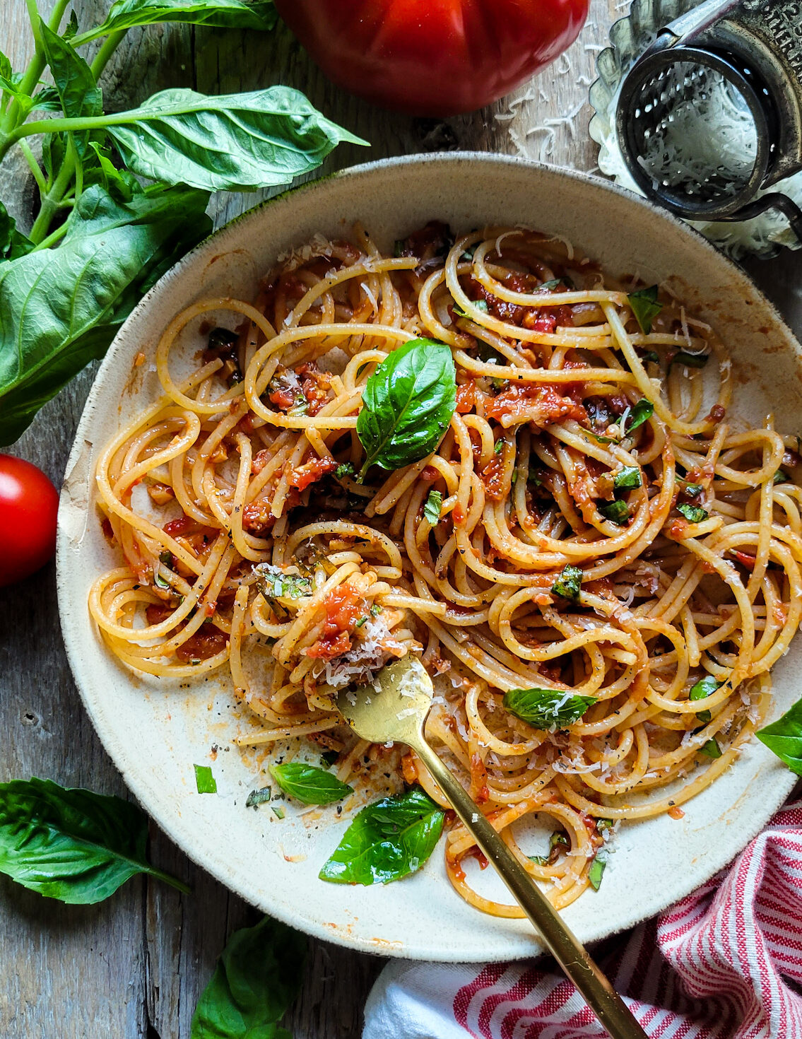A bowl of Spaghetti with fresh Summer Sauce is surrounded by fresh basil, tomatoes and a cheese grater.