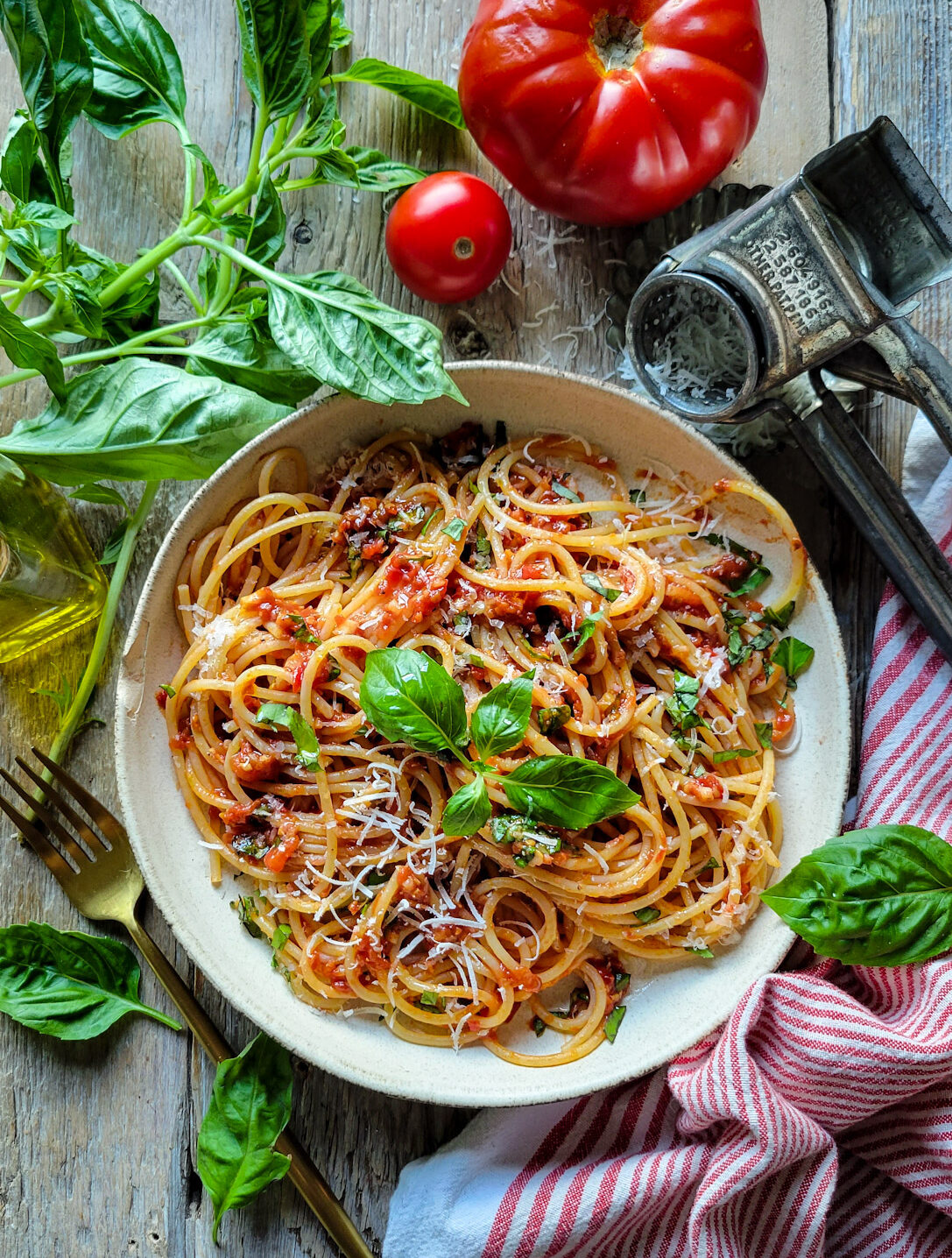 A bowl of Spaghetti with fresh Summer Sauce is surrounded by fresh basil, tomatoes and a cheese grater.